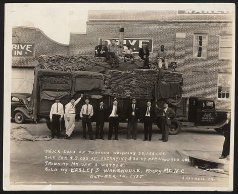 Men standing around and on top of a truck full of bags of tobacco.