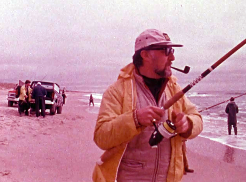 Man in a yellow slicker fishing on the beach, smoking a pipe