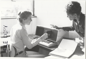 Woman seated before a computer while instructor bends close and points to floppy disk