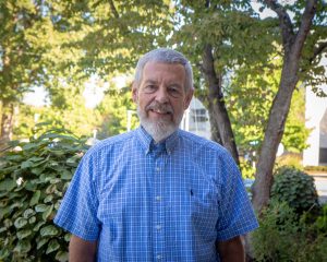Smiling adult with grey beard wearing blue collared shirt standing under trees on a sunny day