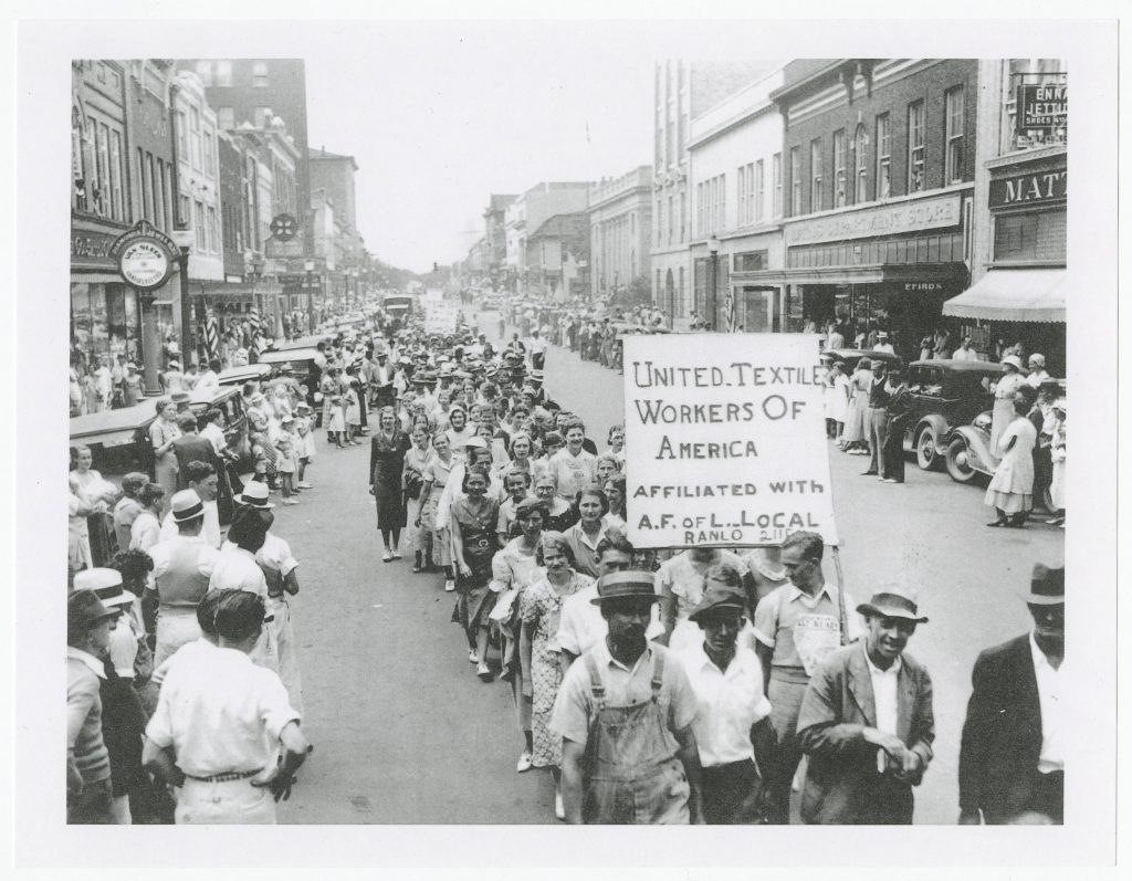 Looking down a main street in Gastonia, NC, showing a line of striking men and women while crowds watch from the sidewalks.