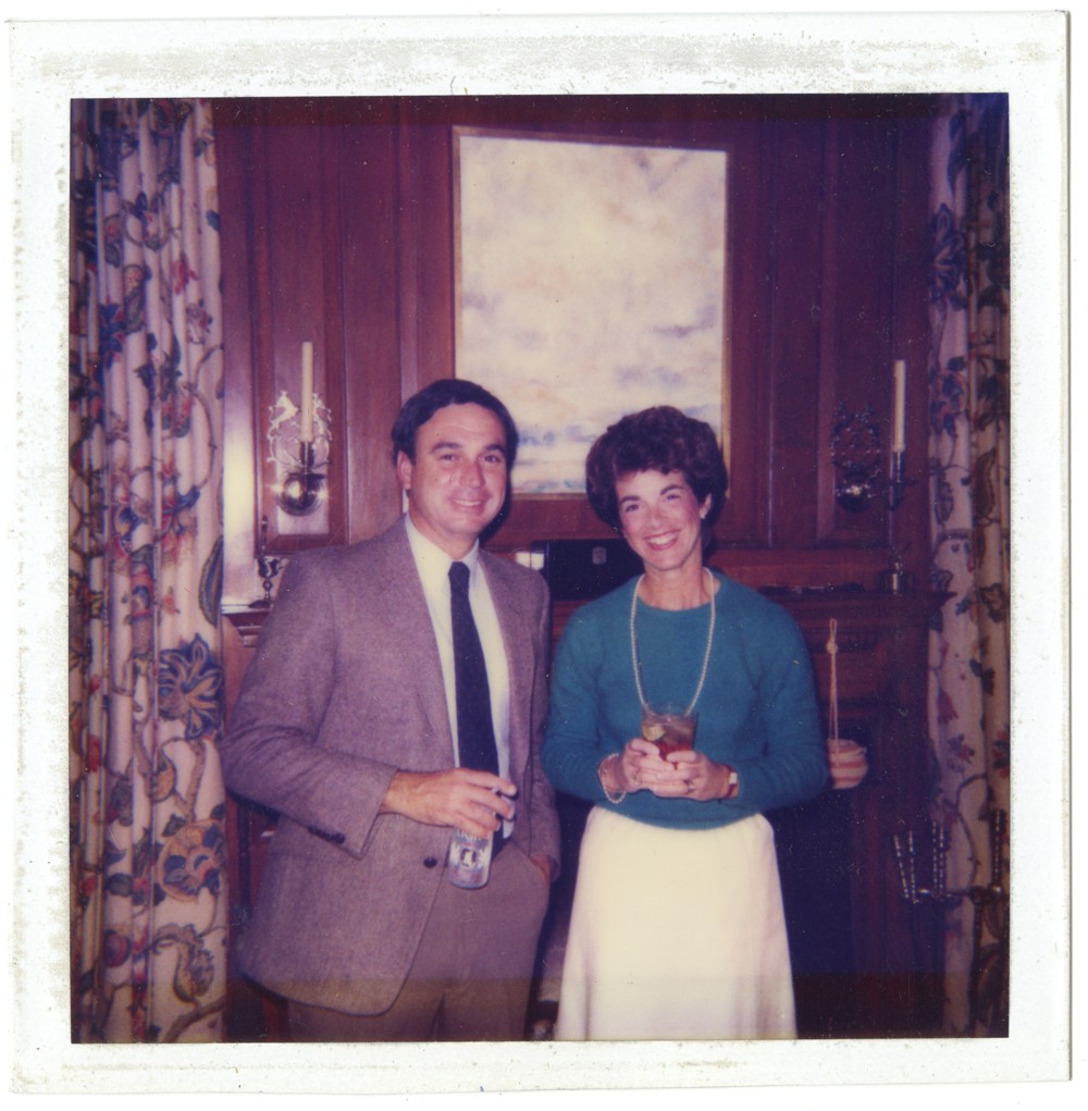 Color photo of two adults standing in front of a fireplace holding beverages.