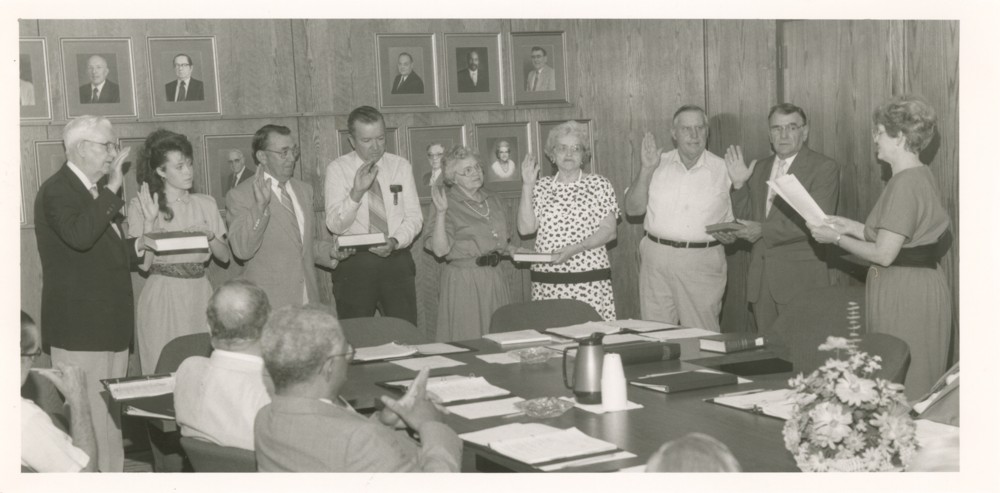 Eight individuals being sworn in to the CCCC Board of Directors. One person stands at the front to do the swearing in while five sitting a table watch on.