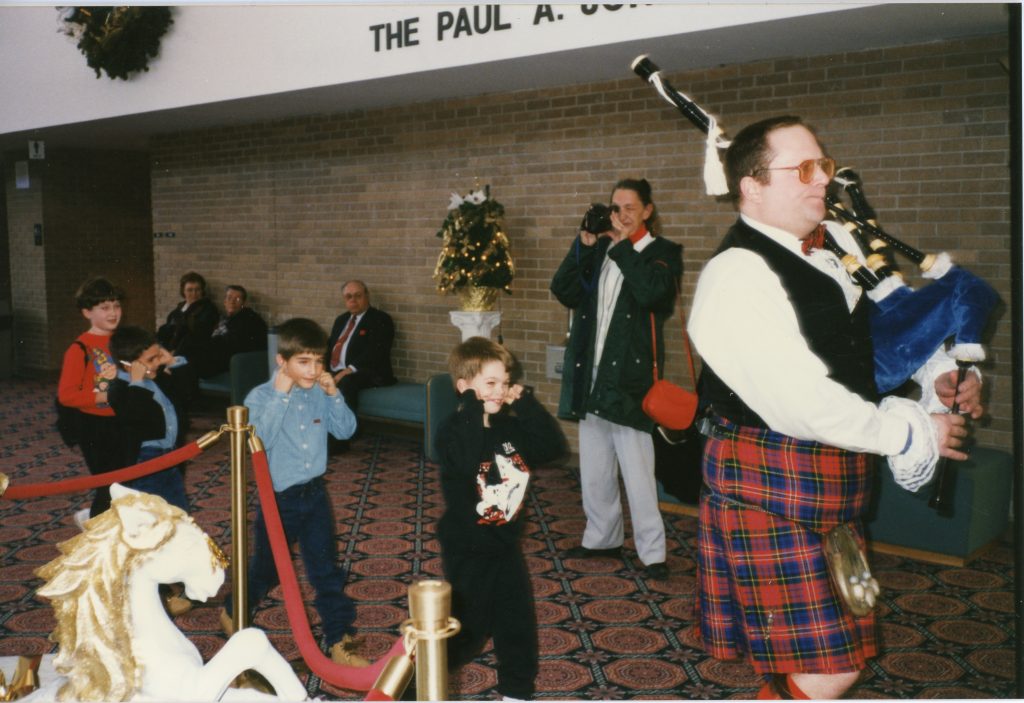 Photo of a bagpipe player with kilt on playing the bagpipe. The musician is walking to the right while three children walk behind the musician, covering their ears.