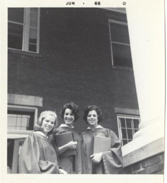 A black and white photo of three smiling students in graduation attire posed in front of a building.