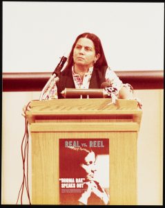 photograph of woman with long hair and a serious face behind a podium with sign that says Real vs Reel
