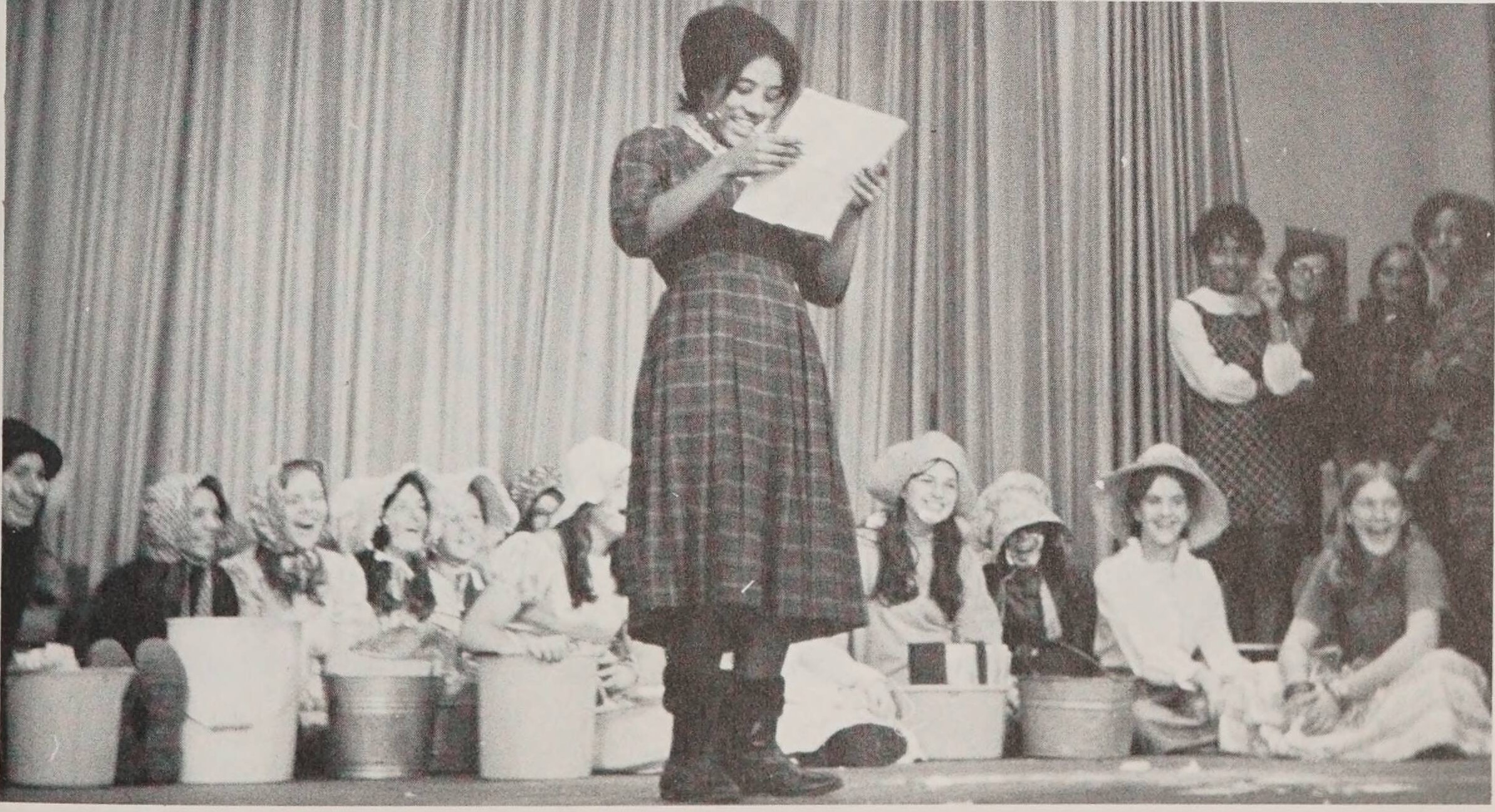 Teenagers on a stage for the F. H. A. initiation. One teen is in the center of the stage holding a piece of paper while others sit behind her with water pales and bonnets on their heads.