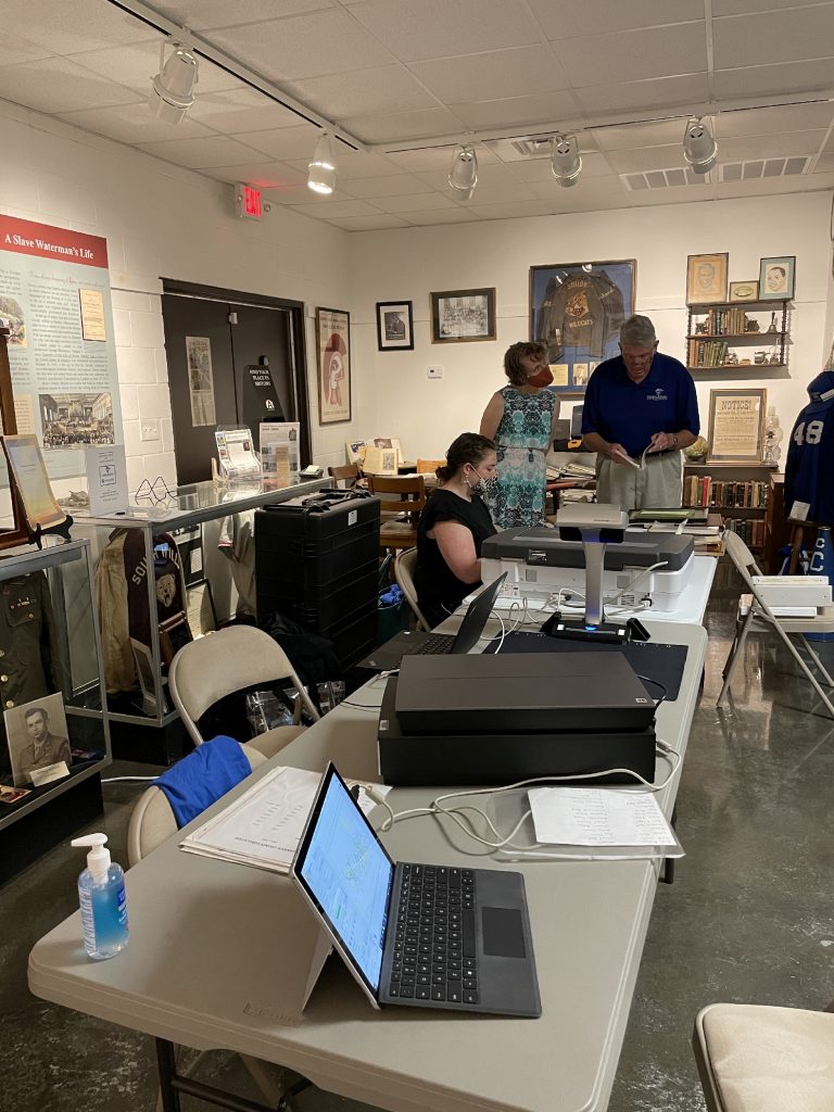 Table with scanners and a laptop inside a brightly lit museum space with one person seated and scanning and two people standing and looking at a book