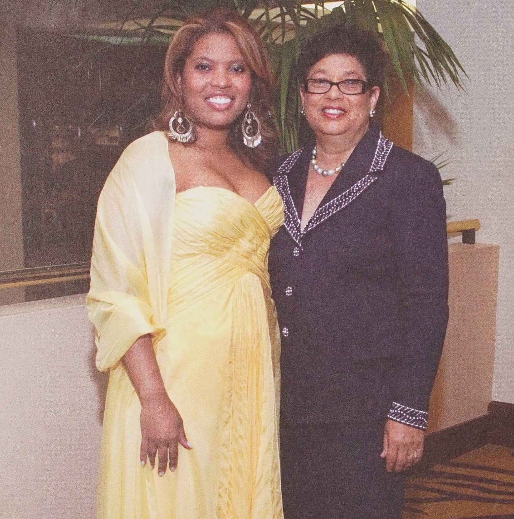 Dr. Dorothy Cowser Yancy and her daughter, Yvonne Cowser Yancy, standing next to each other. The daughter, who is on the left, wears a floor length sunshine yellow gown. The mother, on the right, wears a black blazer with a pattern on the neckline and cuff area with either black pants or a black floor length skirt.