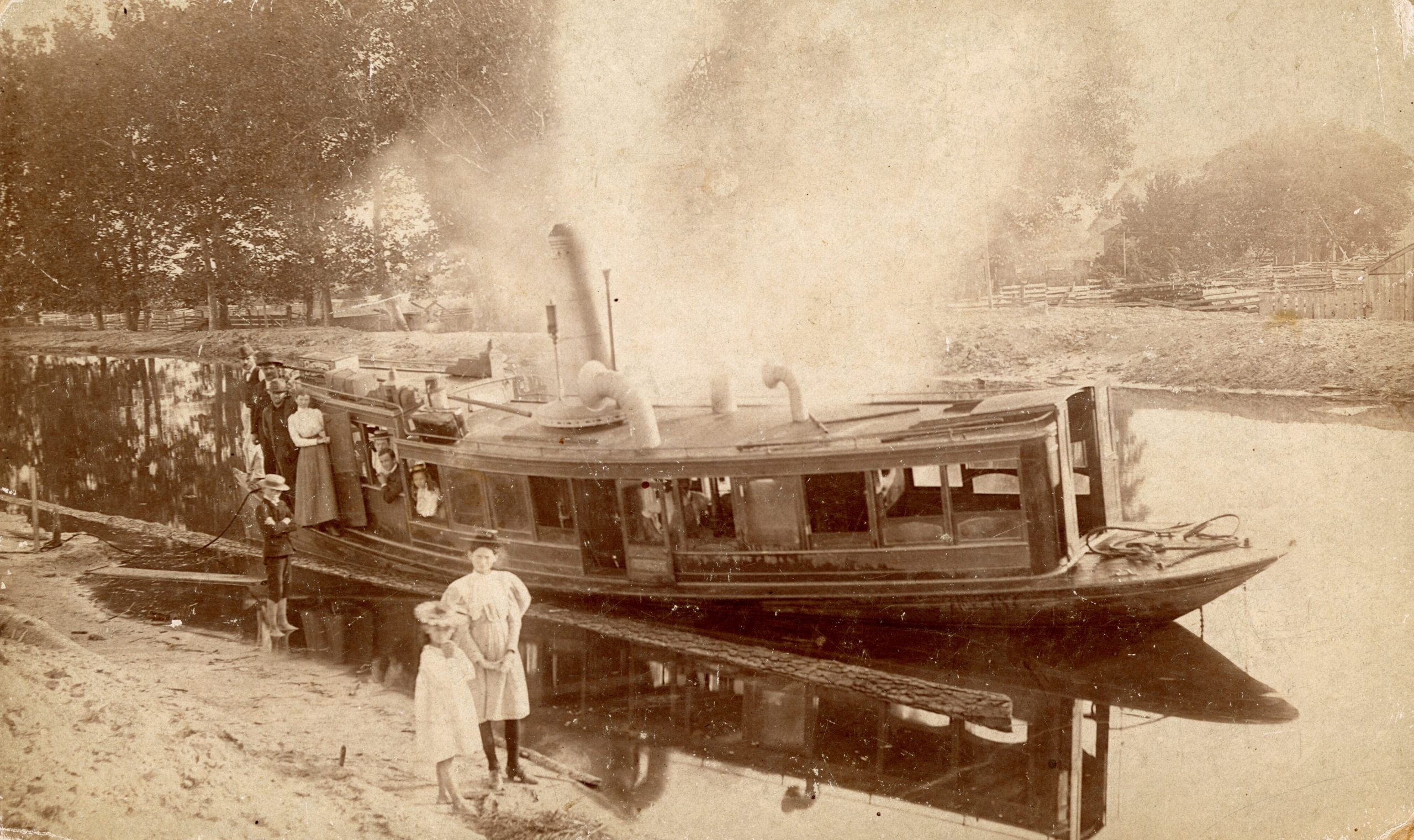 Commercial boat on the side of a canal with several people standing on the boat and two children standing on the shore in front of it.