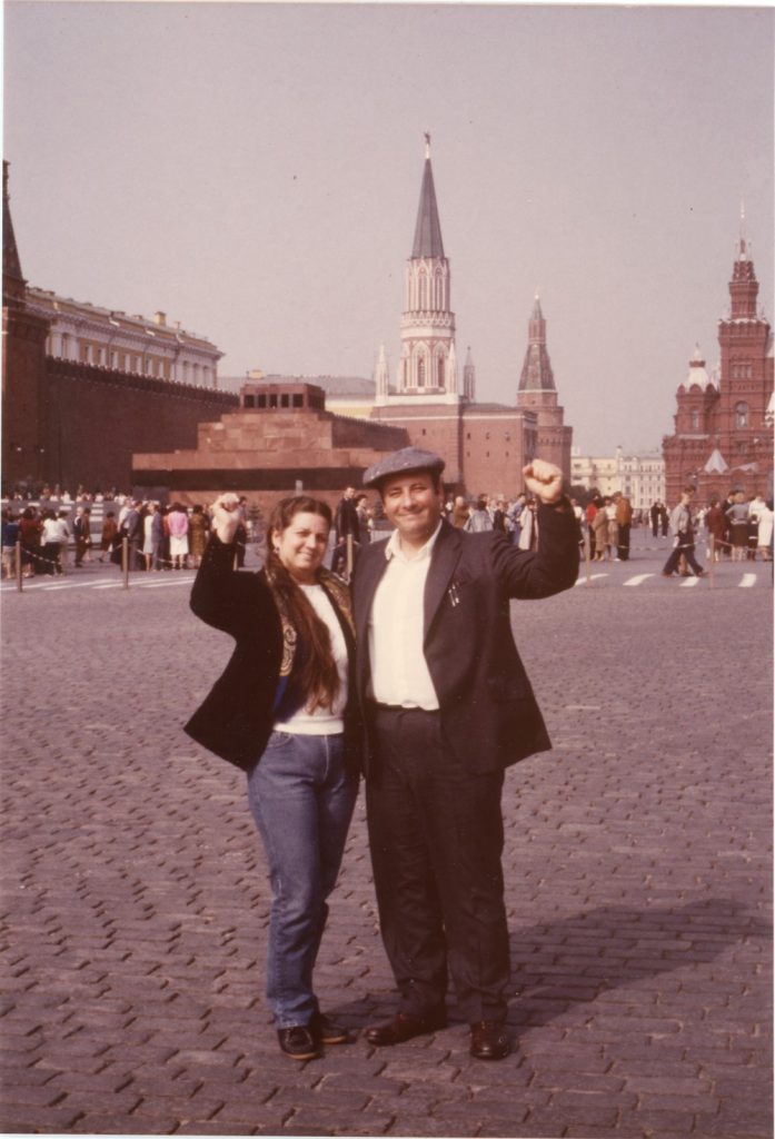 Color photograph of two smiling individuals with raised fists facing camera, Lenin's tomb with line of visitors in the background
