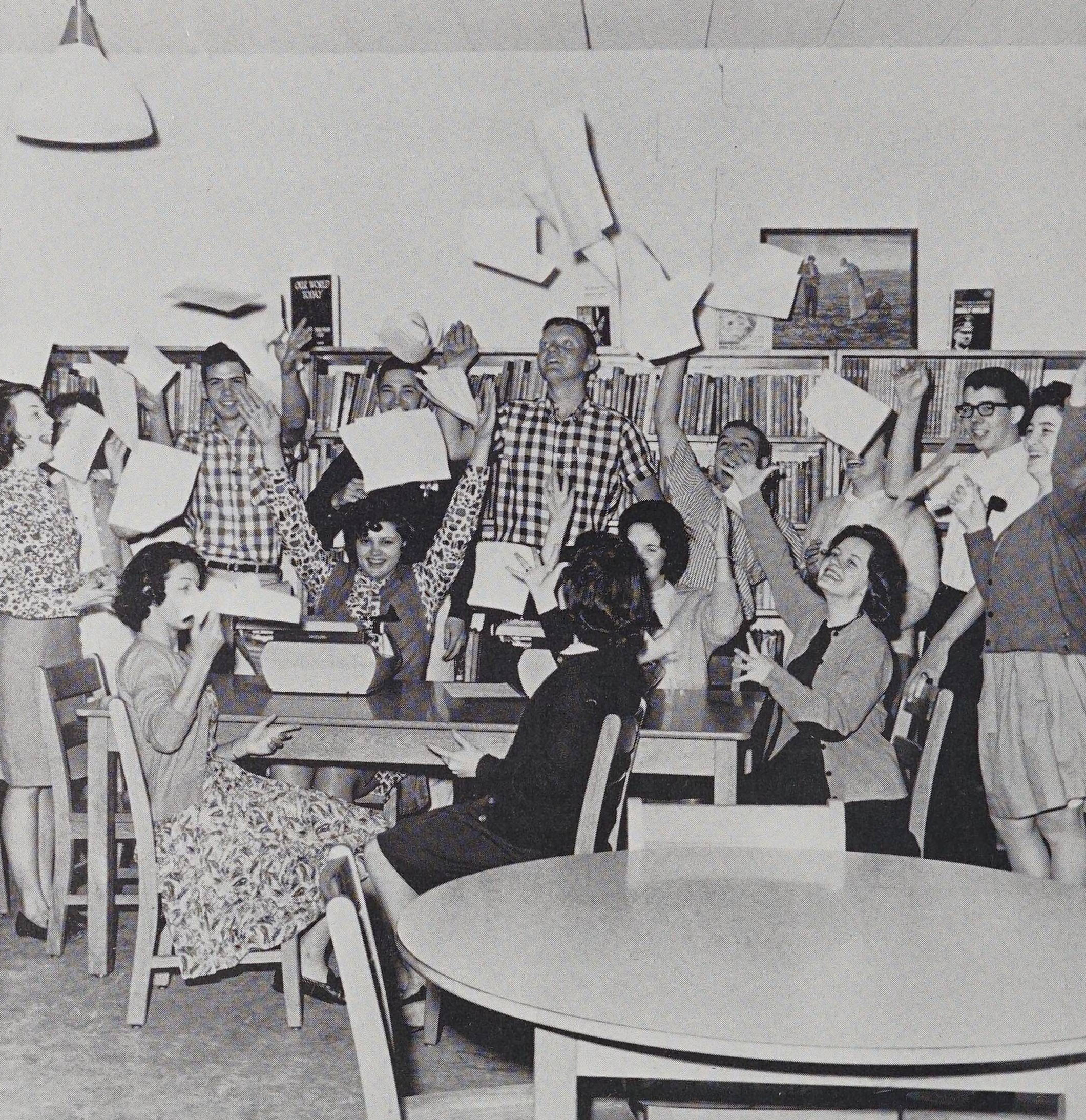Group of students in 1950s fashion in a classroom throwing papers into the air.
