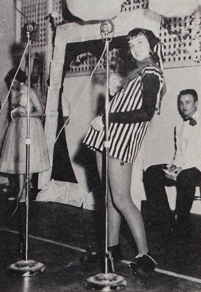Individual dressed in a long striped shirt with bells and stockings stands in front of a microphone looking at the camera.