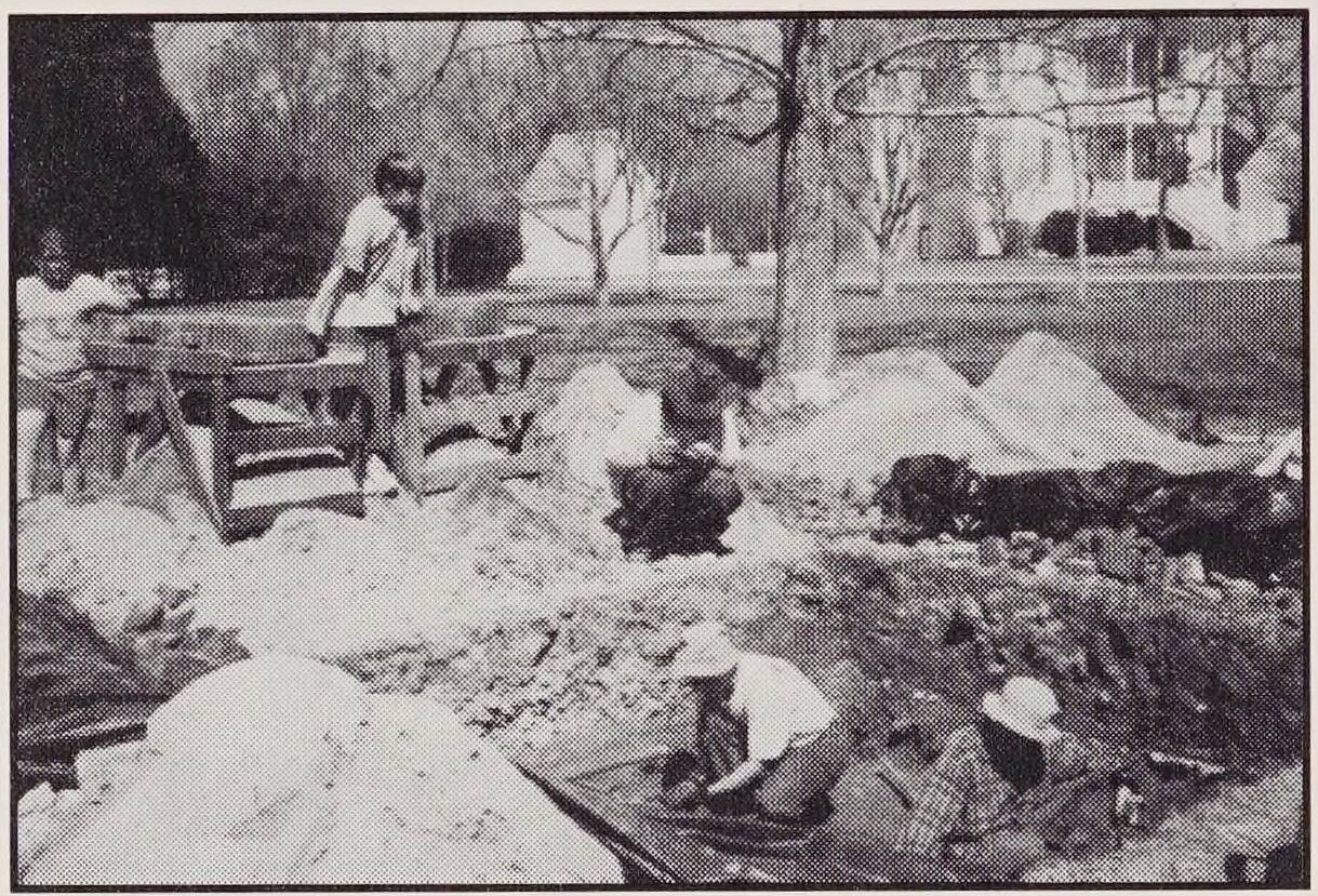 Photograph of five individuals working on excavating at Hope Mansion. There are two people in a pit with several small piles of dirt around them. Another individual crouches near the put. Two other individuals look like they are walking towards the pit.