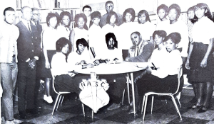 A group of many students gathered closely together. Most are standing in a semi-circle around a table; six are seated at the table.