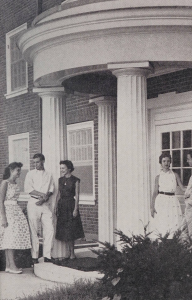 A photo of four Wake Forest College students gathered at the entrance to a dorm. They are standing under a rounded entryway of a brick building with tall, white columns by the door.