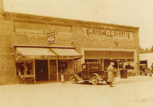 A sepia-toned photo of a one-story brick building. A car is parked in front with a person standing nearby.