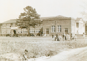 A grayscale photo of a brick building. A tall tree stands beside it. Children are on the grass in the foreground.