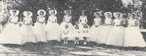 A photograph of 10 women in white dresses standing in a line. Three children stand in front in the middle.