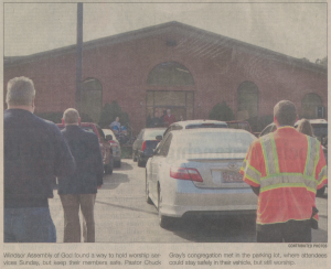 A photo of several people standing in a parking lot outside a church. Their backs are to the camera, and they are watching another person deliver a service.