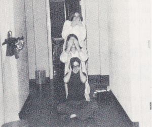 Three students; the one sitting on the floor has hands over ears; the middle one has hands over eyes, and the standing one has hands over mouth.