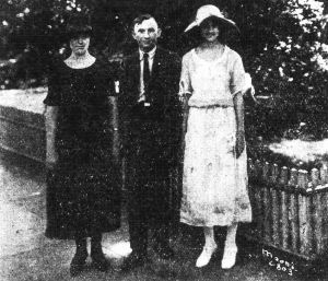 Three people standing together. The person on the left is wearing a black dress and hat; the person in the middle is wearing a suit and tie; the person on the right is in a white dress and hat.