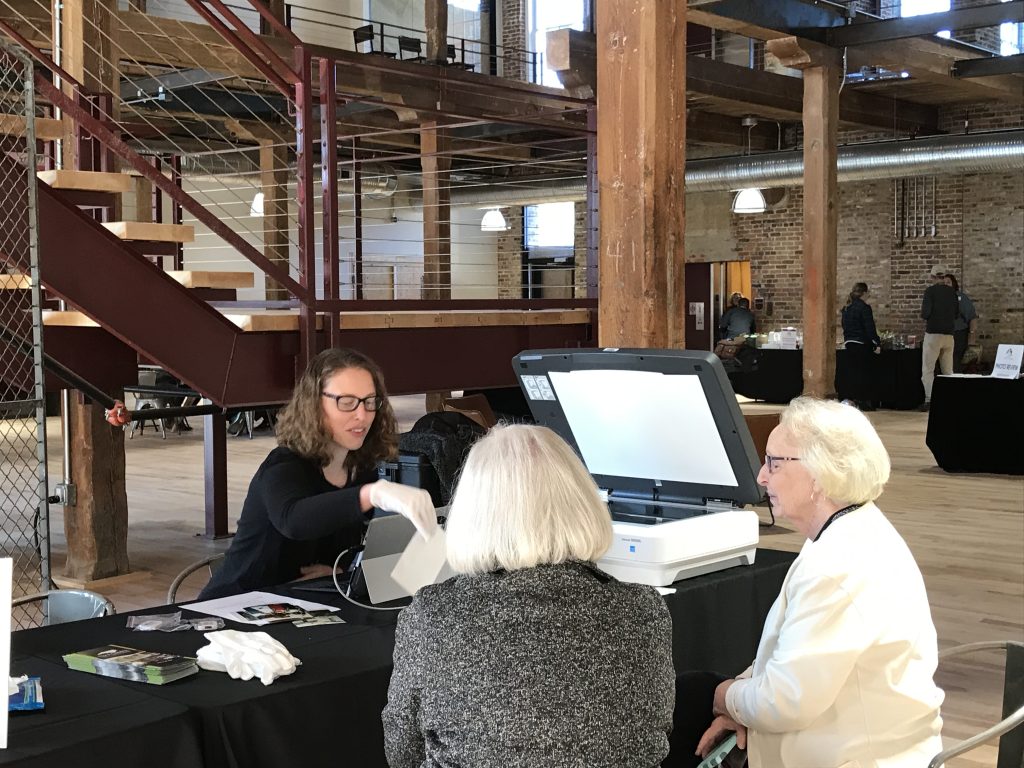 three adults sitting around a table; one with white gloves sits behind a laptop near an open scanner while the other two look on