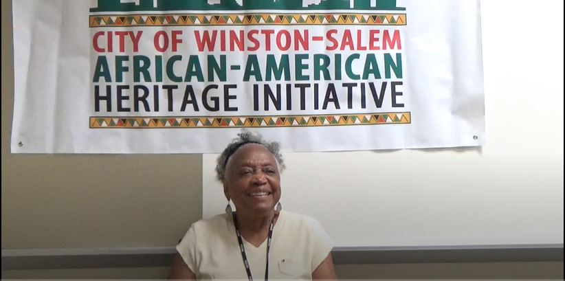 Smiling woman sitting in front of a sign that says City of Winston-Salem African-American Heritage Initiative