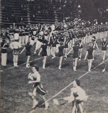 Individuals in marching band and majorette uniforms forming a tunnel structure for football players to run through on a football field.
