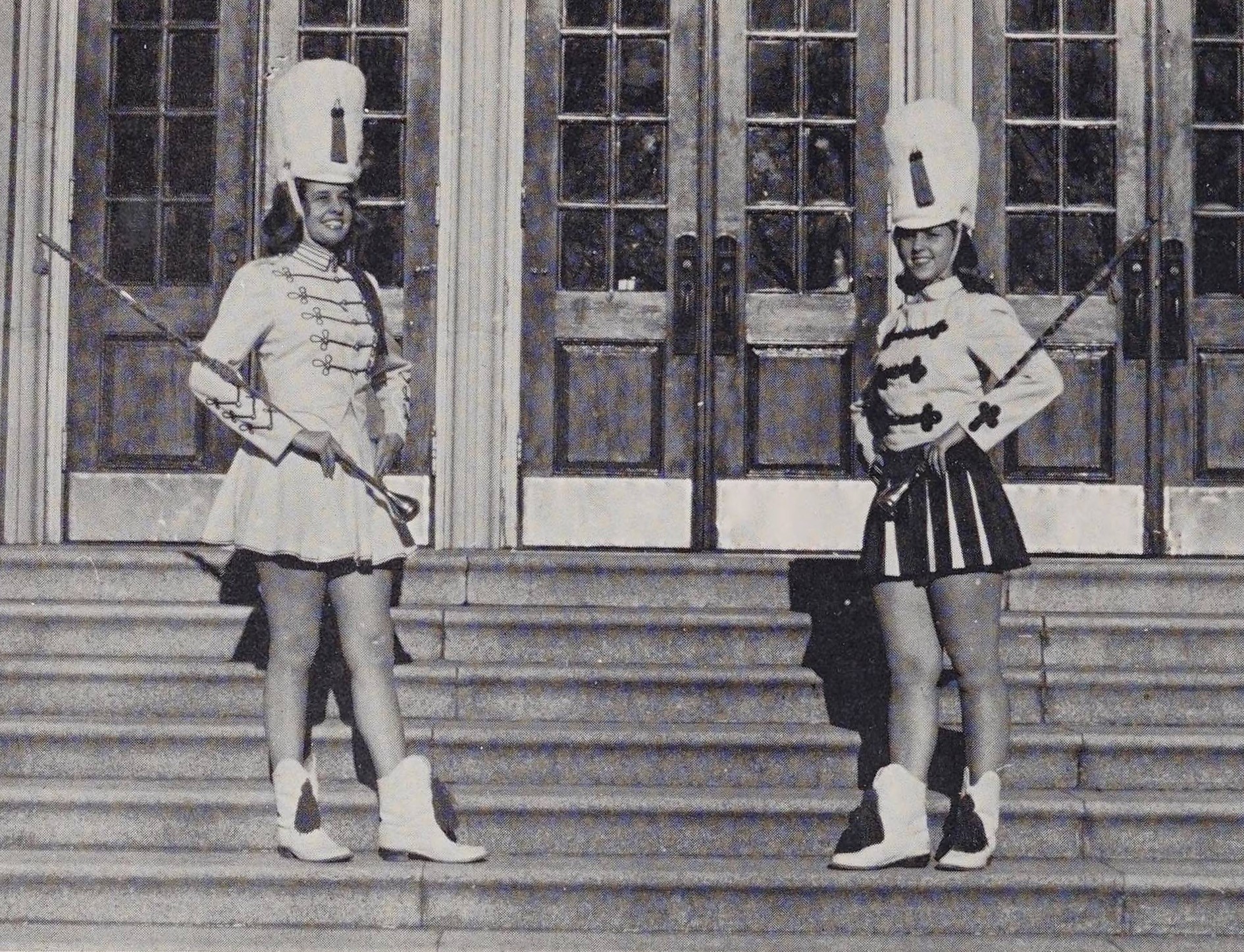 Two individuals standing on steps wearing majorette uniforms with batons in their hands.
