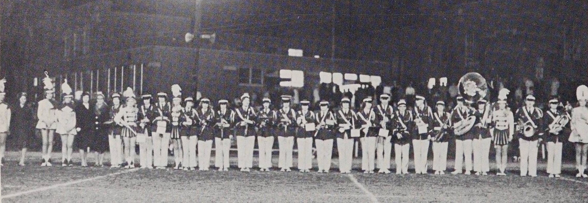 Line of 31 people in majorette and marching band uniforms standing with their various instruments on a football field.