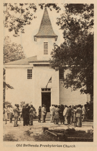 A sepia photo of a white church with a group of people talking in groups out front