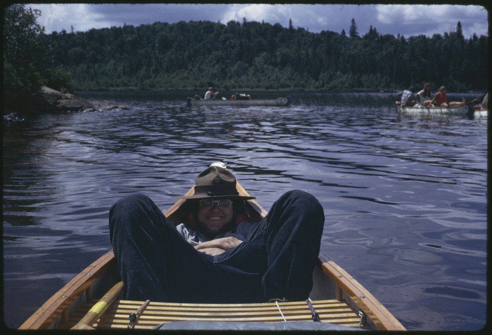Individual in a ranger looking hat and glasses sitting slumped in a canoe. Behind them is a lake with several other canoes filled with people.