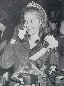 A homecoming queen in a tiara dabs her eye with a tissue as she holds a bouquet of flowers.
