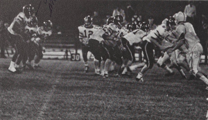 A black-and-white photo of football players clustering together on the field, presumably during a play.