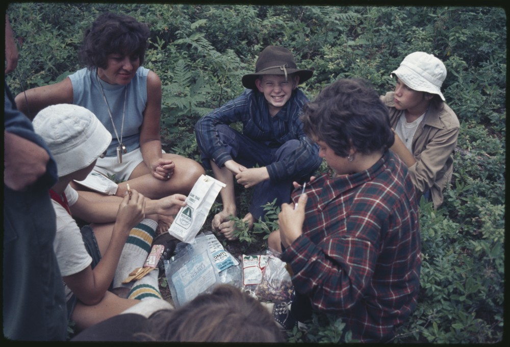 A group of various individuals sitting in a circle on the forest floor looking at different things. One individual wearing a hat is looking at the camera and smiling.