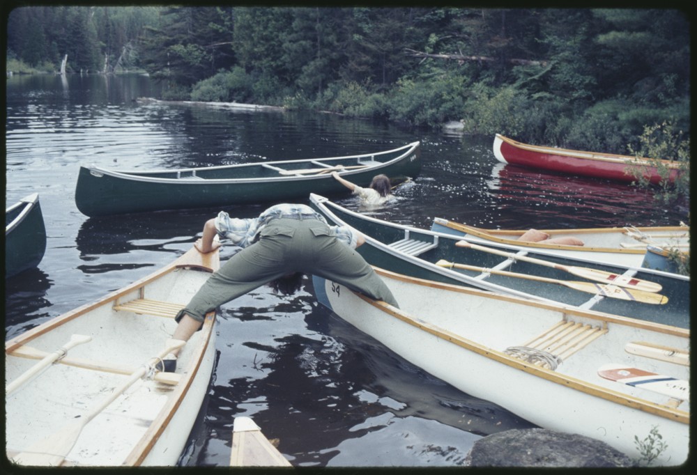 On a lake, an individual stands with each leg in a different boat. The boats seem to be floating away from each other, causing the person to lean over to try to stop themselves from falling.