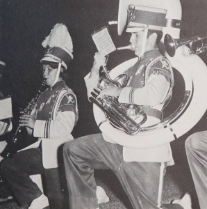 Two band students kneeling. The one in the back (to the left) is playing the clarinet; the one in the front (right) is playing the tuba.