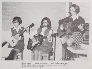 Three students sitting at the front of a classroom. Each of them holds a guitar or similar stringed instrument.