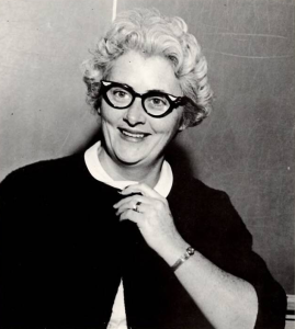 A black-and-white portrait photo of a smiling teacher with short, curly hair and black glasses