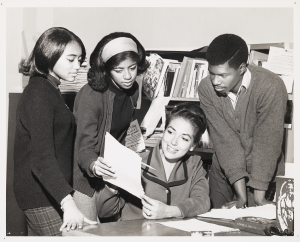 A group of three students gathered around their advisor, seated, all looking at a piece of paper.