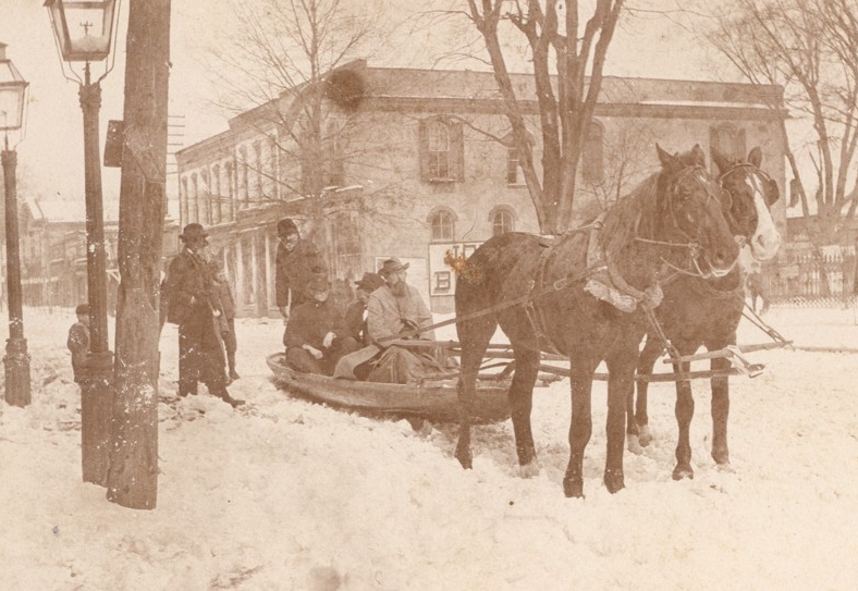 black and white photograph of a snowy downtown with adults sitting in a boat drawn by two horses