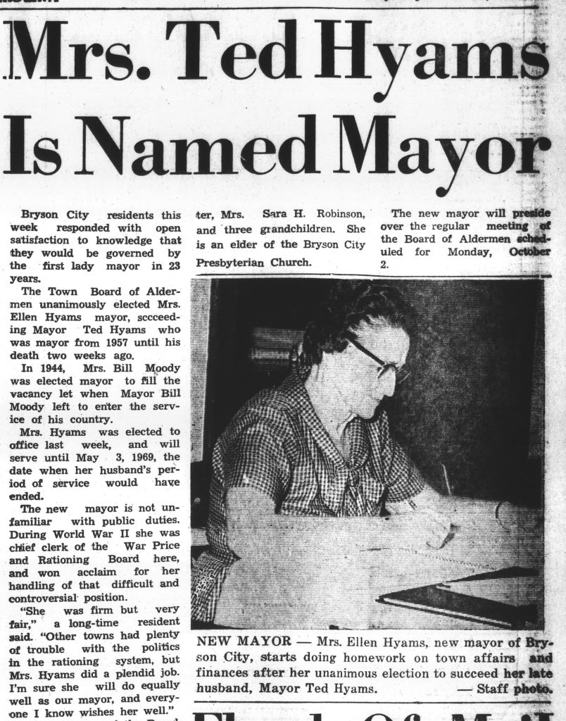 Black and white article text and photo of adult in glasses with checkered dress looking down at paperwork on a desk or table