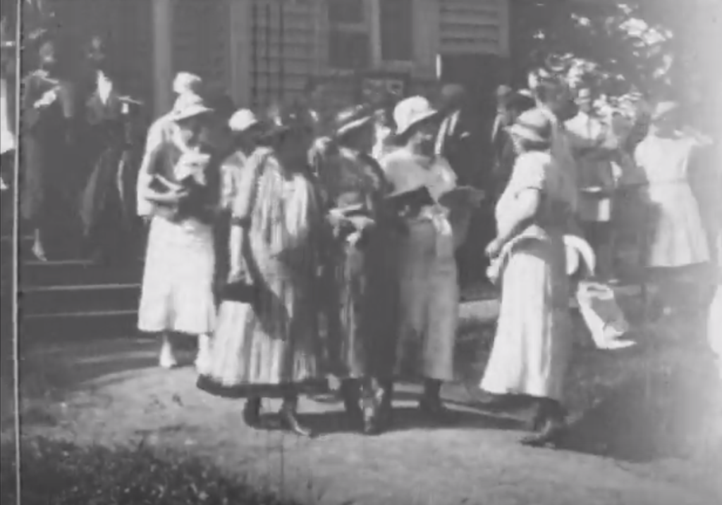 Several adults in dresses and hats talking to one another as they walk out of a house.