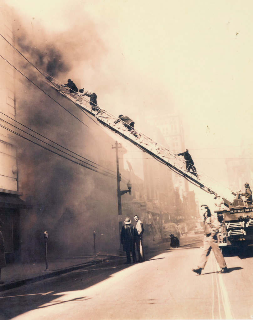 Fire fighters climbing a ladder leaning against a burning building.