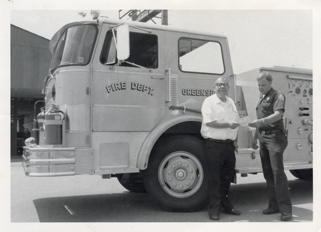 Two adults standing in front of a fire truck. The one on the right is wearing a uniform.