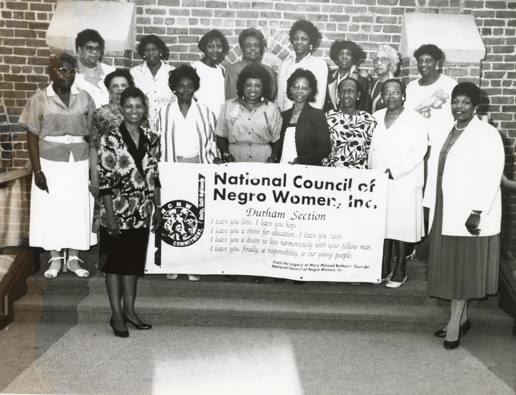 Group of Black women posing on risers. Two of the women are holding a sign that has the council's logo and says "National Council of Negro Women, Inc. Durham Section" and a quote from "From the Legacy of Mary McLeod Bethune."