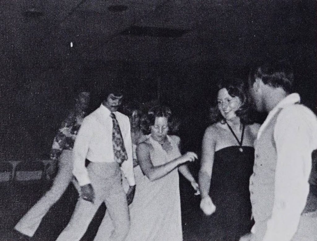 A group of adults in formalwear dancing in a dark room. In the middle are two people stepping to the left of the photo, and on the front left side are two people holding each other.