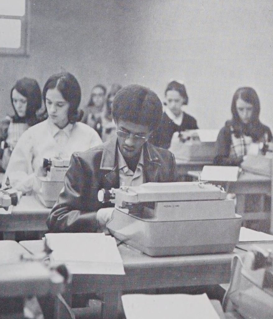 A black-and-white photo of students typing on typewriters at desks in a classroom. In the center of the image is an adult with a short afro and glasses wearing a leather jacket.