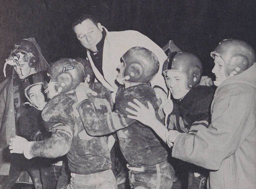 A line of football players in helmets pushing each other playfully while one adult in a white jacket looks at them.